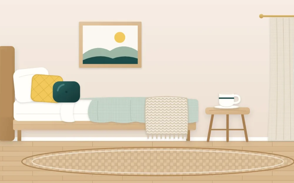 Corner of a bed layered with linen sheets, a chunky knitted throw and two cushions beside a wooden stool, a ceramic cup and a woven rug